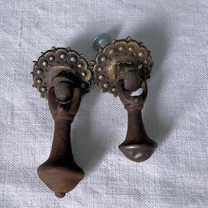 May include: Two antique cabinet door pulls with a gold and brass finish. The pulls have a round decorative top with a ring handle and a long, slender, tapered base.