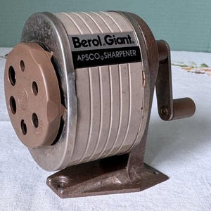 May include: Vintage Berol Giant APSCO sharpener. The pencil sharpener has a brown crank handle and a brown, multi-holed pencil entry. The body is a combination of silver and off-white with the words "Berol Giant APSCO SHARPENER" printed on the front.