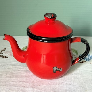 May include: A small red enamel teapot with a black rim and handle. The teapot has a chipped spout and a small dent on the side.