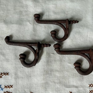 May include: Three rusty metal hooks with a decorative scroll design. The hooks are attached to a white fabric background.