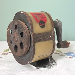 May include: A vintage manual pencil sharpener with a cylindrical body, a dark metal front plate featuring six circular holes, and a side-mounted hand crank. The body displays a red label with the text GIANT and APSCO, and the base is marked MADE IN U.S.A.