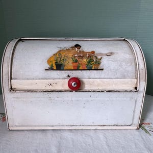 May include: A vintage white metal bread box with a roll-top lid featuring a central graphic of potted plants in various colors. The container has a single round red knob on the front panel and shows signs of wear with visible surface scuffs.
