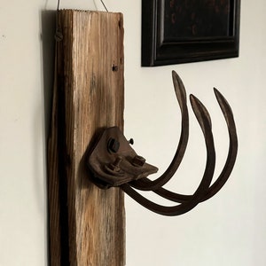 May include: A rustic wall-mounted coat rack made from reclaimed wood and an old metal garden tool. The wood is weathered and has a natural brown color. The metal tool is rusty and has a dark brown color. The rack is hung with a wire loop.