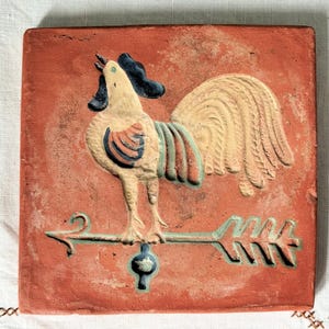 May include: Square, terracotta-colored tile with a raised relief of a rooster weather vane. The rooster is cream-colored with blue and red accents. The arrow points left. A decorative piece for the home.