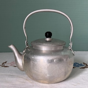 May include: A vintage silver-colored metal teapot with a curved handle and a black knob on the lid. The teapot has a spout for pouring and is likely used for boiling water or brewing tea. The teapot is sitting on a white surface.