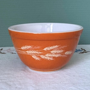 May include: An orange mixing bowl with a white interior and a wheat stalk design. The bowl is round and has a wide rim. The wheat design is in white and is on the exterior of the bowl.