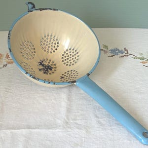 May include: Vintage cream-colored colander with a light blue rim and handle. The colander has numerous small holes for draining and a small loop on the side. A classic kitchen utensil.