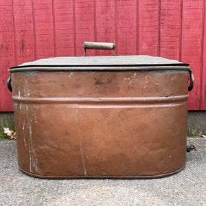 May include: A large, oval-shaped copper wash tub featuring a flat lid with a central wooden handle. The exterior surface shows a weathered, metallic patina with visible signs of wear, including light scuffs and faint white paint splatters.