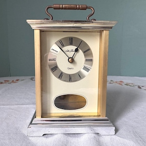 May include: A rectangular mantel clock with a metallic gold and silver frame, a top handle, and a cream-colored face featuring black Roman numerals and the text Seth Thomas Quartz. A small oval window is positioned below the clock face.