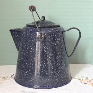 May include: A dark blue enamel coffee pot with a white speckled pattern. The pot features a wide base, a tapered body, a side handle, a spout, and a removable lid with a metal bail handle that has a small wooden grip at the top center.