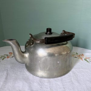 Peut inclure: Une théière en aluminium argenté avec une poignée en plastique noir et un couvercle argenté. La théière a un bec verseur sur le côté et un petit bouton sur le dessus.