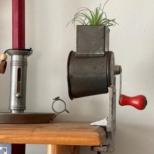May include: A vintage metal food grinder with a red crank handle and a small potted air plant on top. The grinder is mounted on a wooden shelf.