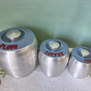 Peut inclure: Trois boîtes en argent avec des couvercles bleus et des inscriptions rouges. Les boîtes sont étiquetées "Farine", "Café" et "Thé".