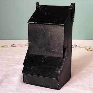 May include: A black wooden bird feeder with a hinged door. The feeder has a simple design and is made of unfinished wood.