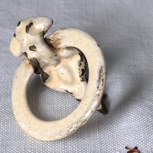 May include: An antique, off-white metal drawer pull with a unique, abstract design. The pull features a circular handle and a decorative element with dark brown spots. The paint is chipped, giving it a vintage appearance.