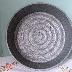 May include: A galvanized metal tub with a circular design. The tub is gray and has a textured surface. The tub's interior features concentric circles, and the rim is slightly darker. This vintage tub is suitable for various uses.
