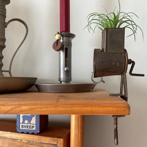 May include: A vintage metal grinder with a wooden handle, attached to a wooden table. A small potted plant sits on top of the grinder. The grinder is rusty and has a dark brown finish. The table is made of light brown wood. The plant is green and has long, thin leaves.