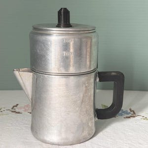 May include: A vintage aluminum coffee percolator with a black handle and lid knob. The percolator has a classic design with a spout and a measurement indicator for 2 cups. It sits on a floral patterned tablecloth.