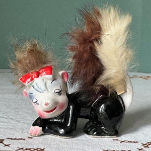 May include: A ceramic figurine of a skunk with a black and white fur tail, wearing a red bow. The skunk has a white face with black eyes and a pink nose.