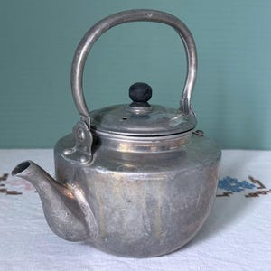 May include: A vintage silver-toned metal teapot with a curved handle and a spout. The teapot has a lid with a black knob. The teapot is sitting on a white tablecloth with floral embroidery.