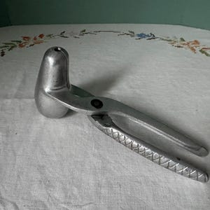 May include: A vintage, silver-colored citrus juicer. The juicer has a rounded pressing head and long handles with a textured grip. The tool is designed to extract juice from lemons, limes, and other citrus fruits. It is made of metal and sits on a white surface.