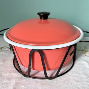 May include: A vintage pink enamelware casserole dish with a white rim and a black knob on the lid. The dish is sitting on a black metal stand.