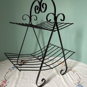May include: Black metal magazine rack with a two-tiered design. The rack features a curved base and decorative scrollwork at the top. The rack is made of metal and has a vintage aesthetic.