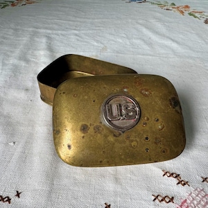 May include: A brass box with a lid. The lid has a circular emblem with the letters "US" in the center. The box is tarnished and has a worn appearance.