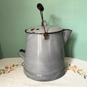 May include: A vintage gray enamelware coffee pot with a metal handle and a spout. The pot has a worn, rustic look and is sitting on a white tablecloth with a floral pattern.