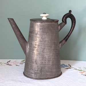 May include: A tall, tapered metal coffee pot with a weathered gray finish. It features a long, angled spout, a dark brown curved handle, and a flat lid topped with a small, white ceramic knob. The body has a textured, matte appearance.