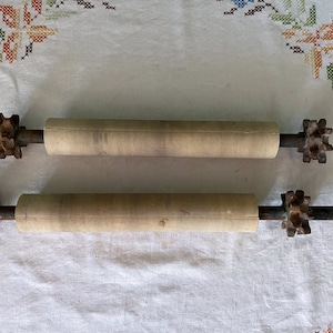 May include: Two wooden rollers with metal ends. The rollers are covered in a light brown material and have a rough texture. The metal ends are rusty and have a star-shaped design.