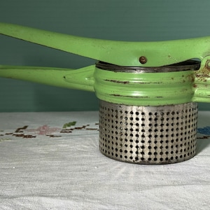 May include: A vintage potato ricer with a bright green handle and a perforated metal base. The ricer has a lever-style design for easy pressing. The metal shows signs of wear, suggesting it's an older kitchen tool.