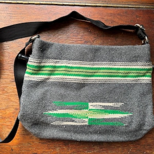May include: Grey woven crossbody bag with green and white stripes and a geometric design. The bag has a black adjustable strap.