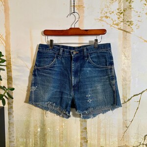 以下が含まれることがあります： フリンジの付いた青いデニムのショートパンツ。ハイウエストで、いくつかの穴があいてダメージ加工が施されています。