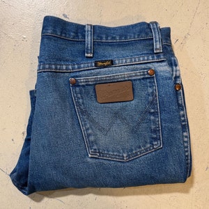 Pu&ograve; includere: Un paio di jeans blu in denim piegati a met&agrave;. I jeans hanno una toppa in pelle marrone sulla tasca posteriore con la scritta "Wrangler" stampata su di essa.