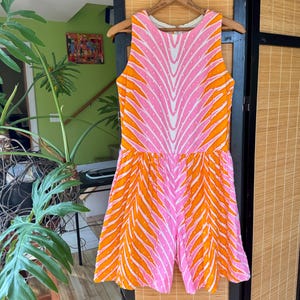 May include: A sleeveless romper featuring a vibrant pattern of alternating pink and orange vertical stripes with white accents. The garment has a round neckline, a fitted bodice, and a flared bottom section, all displayed on a wooden hanger.