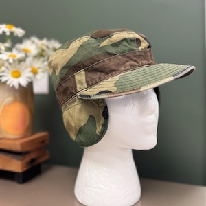 May include: A camouflage print cadet cap featuring a flat top, a stiffened brim, and fold-down ear flaps. The hat is constructed from a durable fabric with a pattern of green, brown, and tan patches, designed to provide coverage for the ears.