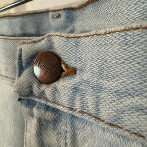 May include: Close-up of a light blue denim button with the word "Wrangler" embossed in brown on the button.