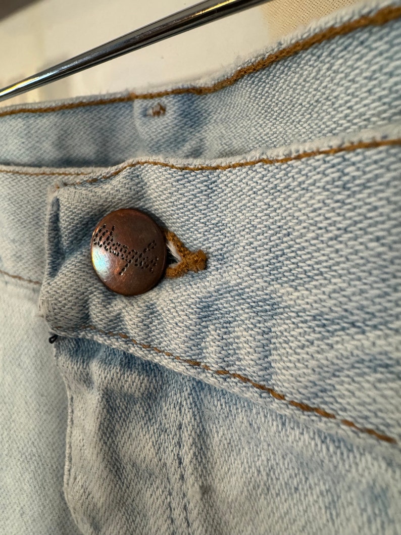 May include: Close-up of a light blue denim button with the word "Wrangler" embossed in brown on the button.