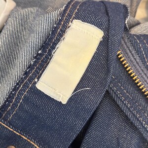 May include: Close-up of a dark blue denim jacket with a white label sewn on the collar. The label has a stitched border and is blank.