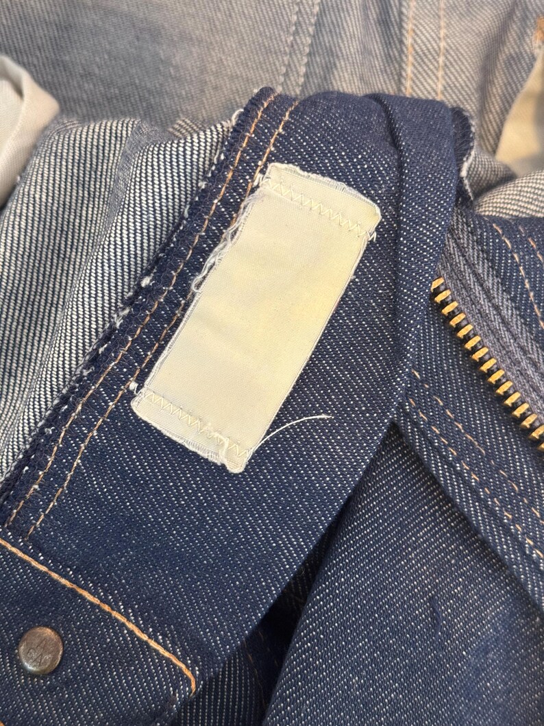 May include: Close-up of a dark blue denim jacket with a white label sewn on the collar. The label has a stitched border and is blank.