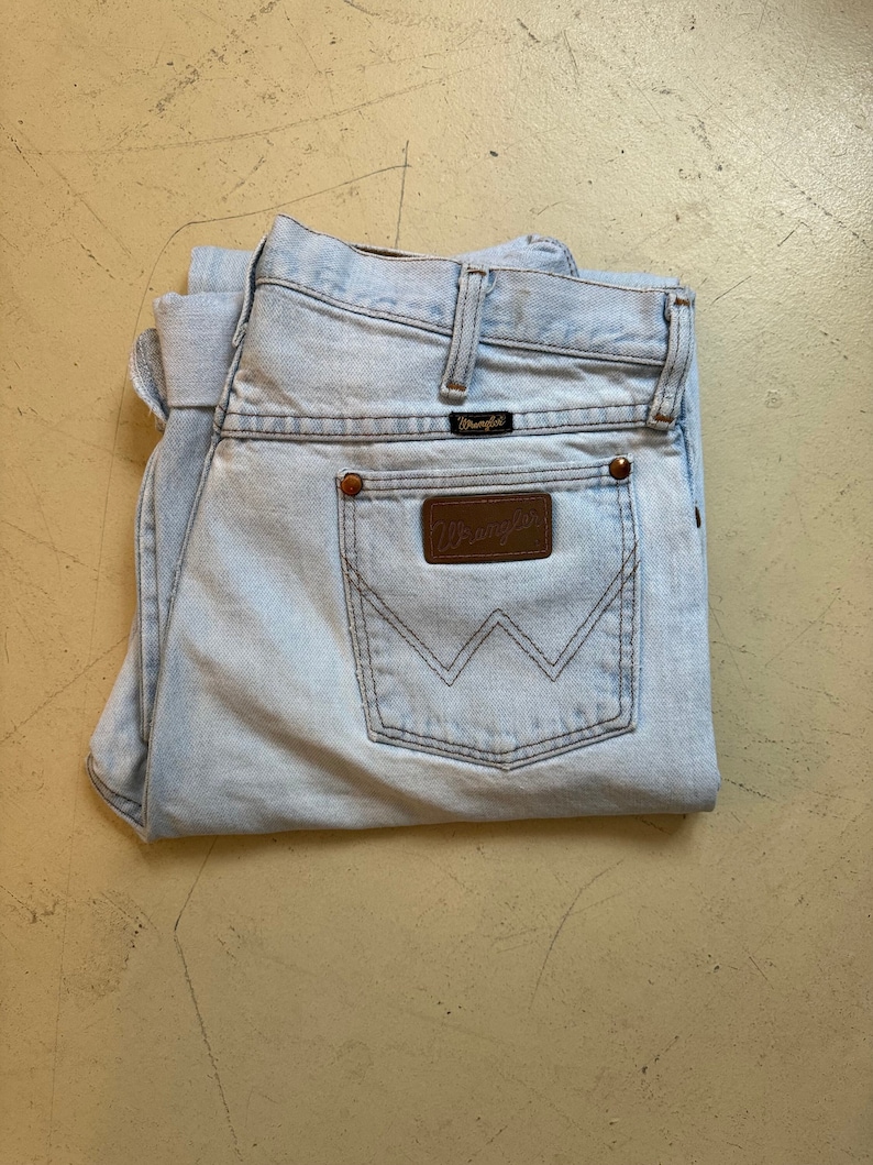 May include: A pair of light blue denim jeans with a brown leather Wrangler patch on the back pocket.