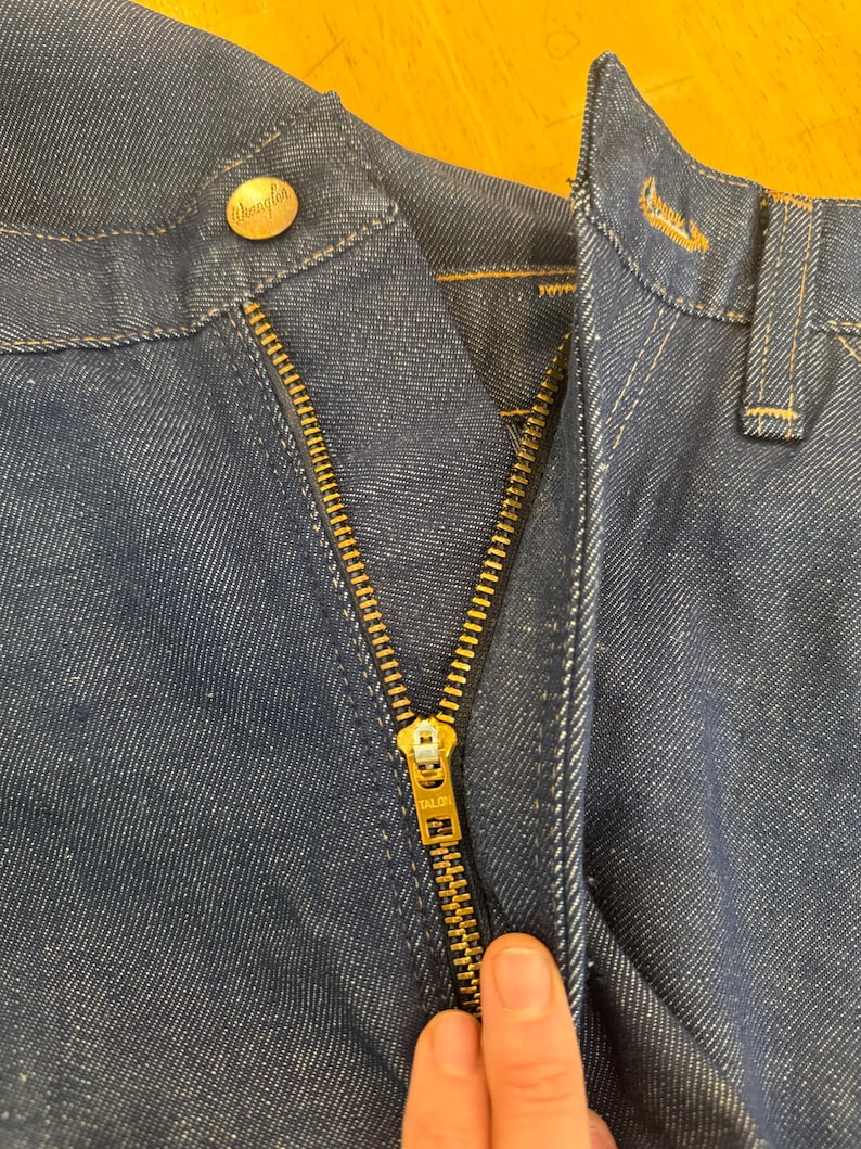 May include: Close-up of a blue denim zipper fly with a gold zipper and a "Wrangler" button. The zipper has a "TALON" label.