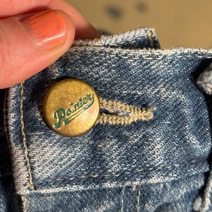 Könnte beinhalten: Nahaufnahme eines goldenen Knopfes mit dem Wort "Painter" in grüner Schrift auf einer blauen Jeans.