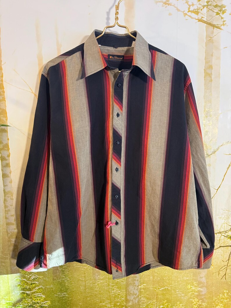 May include: A long-sleeved button-down shirt with vertical stripes in shades of black, red, orange, and gray. The shirt has a pointed collar and a button-up front.