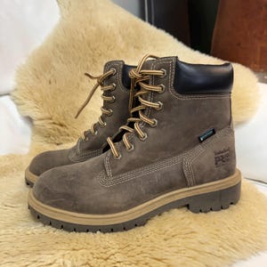 Puede incluir: Una bota de trabajo de cuero marrón con cuello acolchado negro, cordones a rayas marrones y beige, y una suela de goma gruesa de color beige. El lateral presenta un logotipo Timberland PRO y una etiqueta negra con la palabra waterproof.
