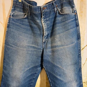 Pu&ograve; includere: Un paio di jeans blu denim con chiusura a bottone e cinque tasche. I jeans sono usurati e sbiaditi, con un aspetto vintage.