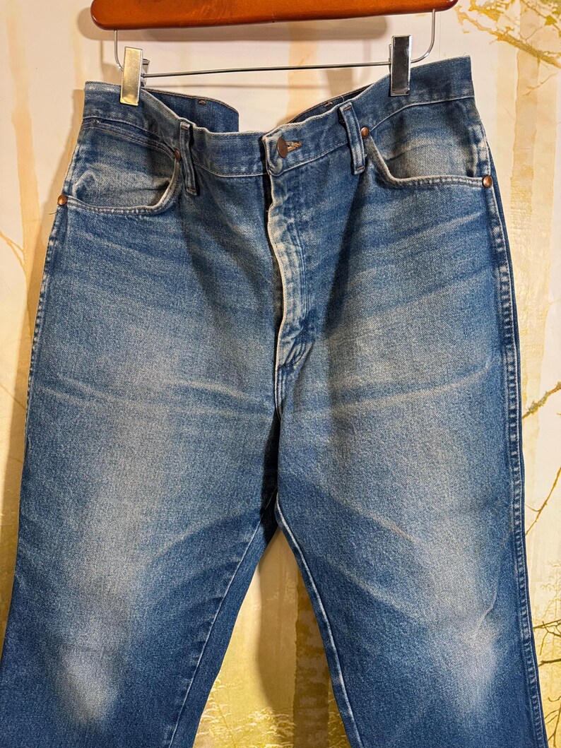 Pu&ograve; includere: Un paio di jeans blu denim con chiusura a bottone e cinque tasche. I jeans sono usurati e sbiaditi, con un aspetto vintage.