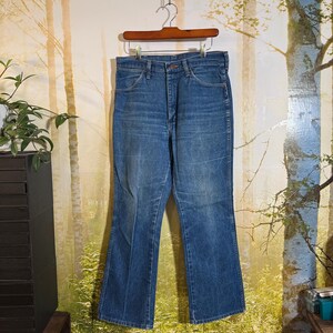 Puede incluir: Un par de pantalones vaqueros azules de mezclilla con abertura de pierna acampanada. Los vaqueros est&aacute;n colgados de una percha de madera sobre un fondo de escena de bosque.