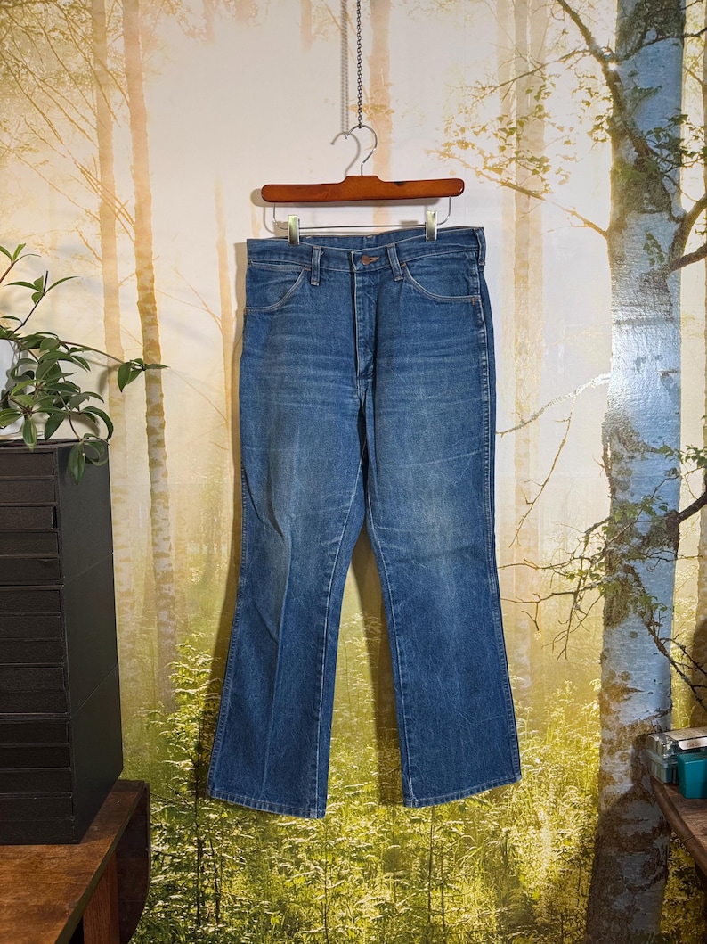 Puede incluir: Un par de pantalones vaqueros azules de mezclilla con abertura de pierna acampanada. Los vaqueros est&aacute;n colgados de una percha de madera sobre un fondo de escena de bosque.
